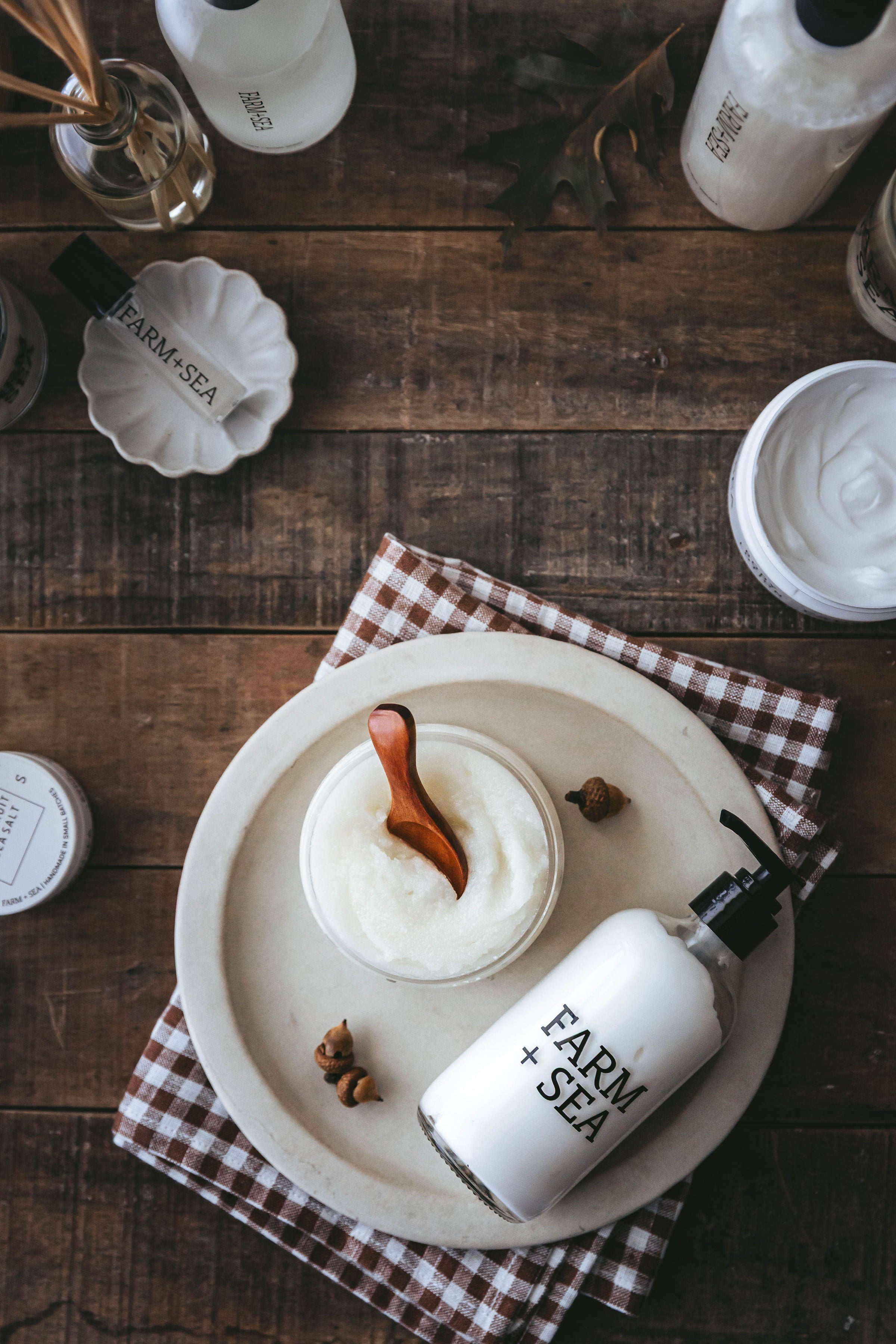A styled flat-lay of a white Farm + Sea body lotion bottle next to a jar of white body cream on a ceramic plate, set on a rustic wooden table with a gingham napkin.