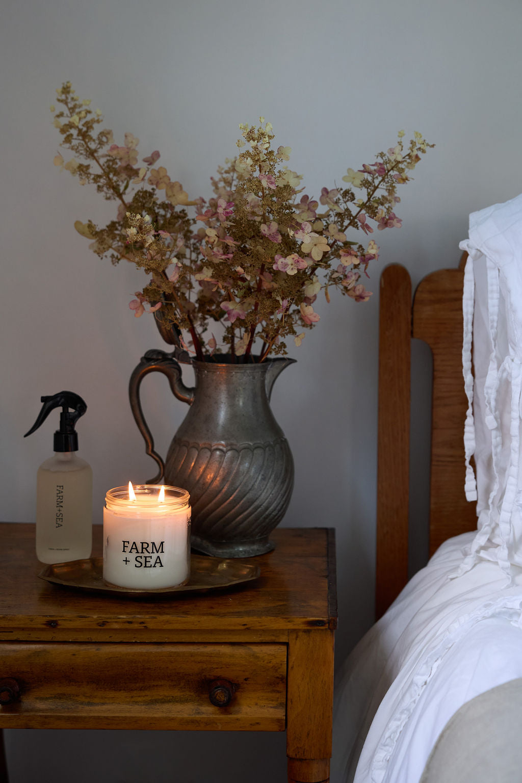 A lit white candle labeled 'FARM + SEA' sits on a wooden bedside table next to a glass Farm + Sea Linen + Room spray bottle and a large rustic metal pitcher filled with dried pink and beige flowers, with a bed visible on the right.