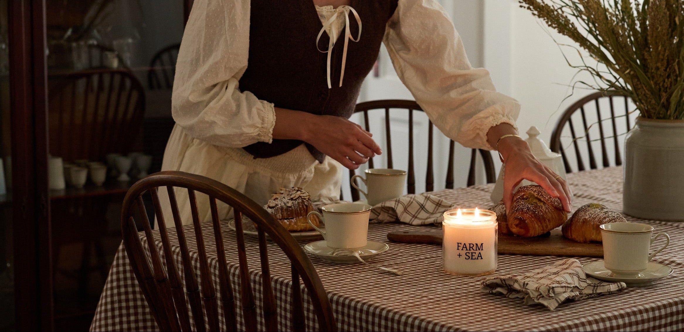 A woman in a vintage-style white blouse and dark brown vest stands at a dining table set with a checkered tablecloth, Farm + Sea candles, and chairs in a warmly lit room.