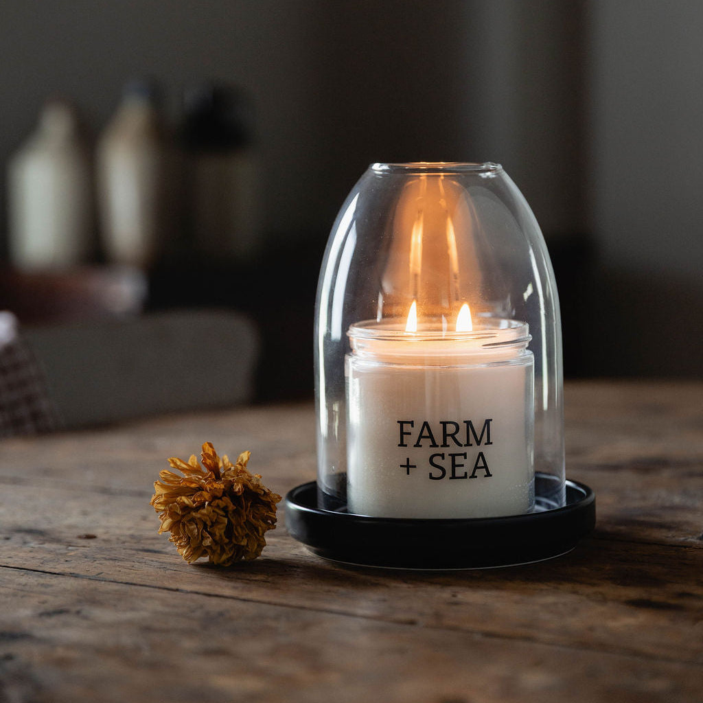 A lit Farm + Sea large, Classic Candle in a hurricane bell jar on a black dish, is placed on a rustic wooden table. The candle is printed with the text 'FARM + SEA' in black.