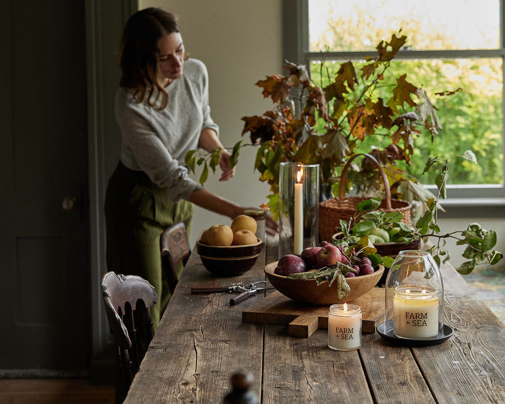A woman in a white sweater and green skirt arranges plants and Farm + Sea Classic Candles on a rustic wooden dining table in a cozy, sunlit room with a black chandelier overhead.