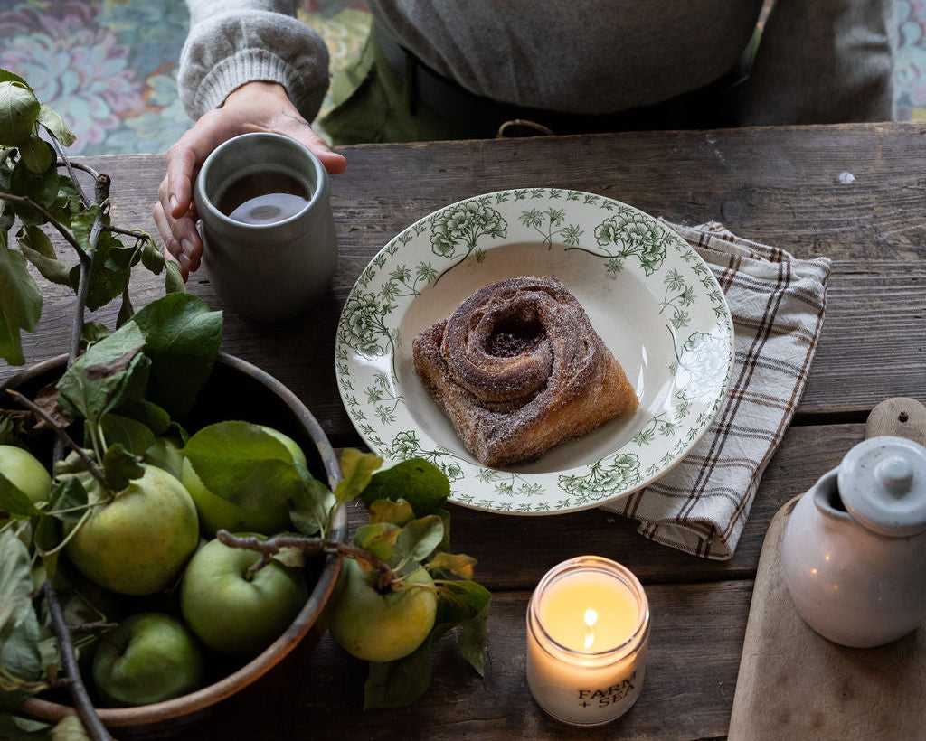A cozy fall tabletop scene with two pastries on green-and-white patterned plates, lit Farm and Sea candles, a bowl of green apples, and part of a person holding a coffew cup at the top of the frame.