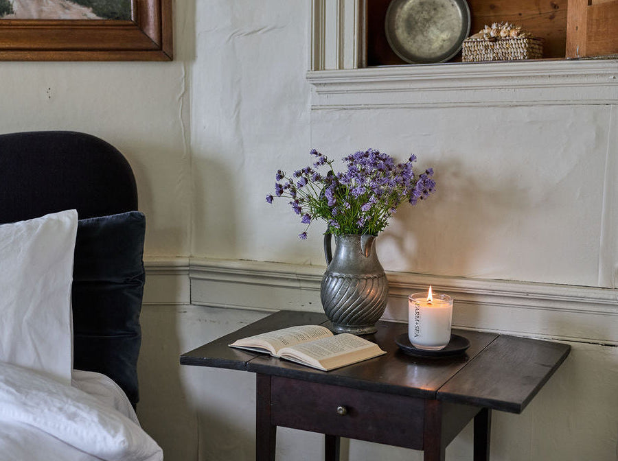 Farm + Sea tumbler candle on a wooden night stand, next to a vase full of purple flowers and an open book