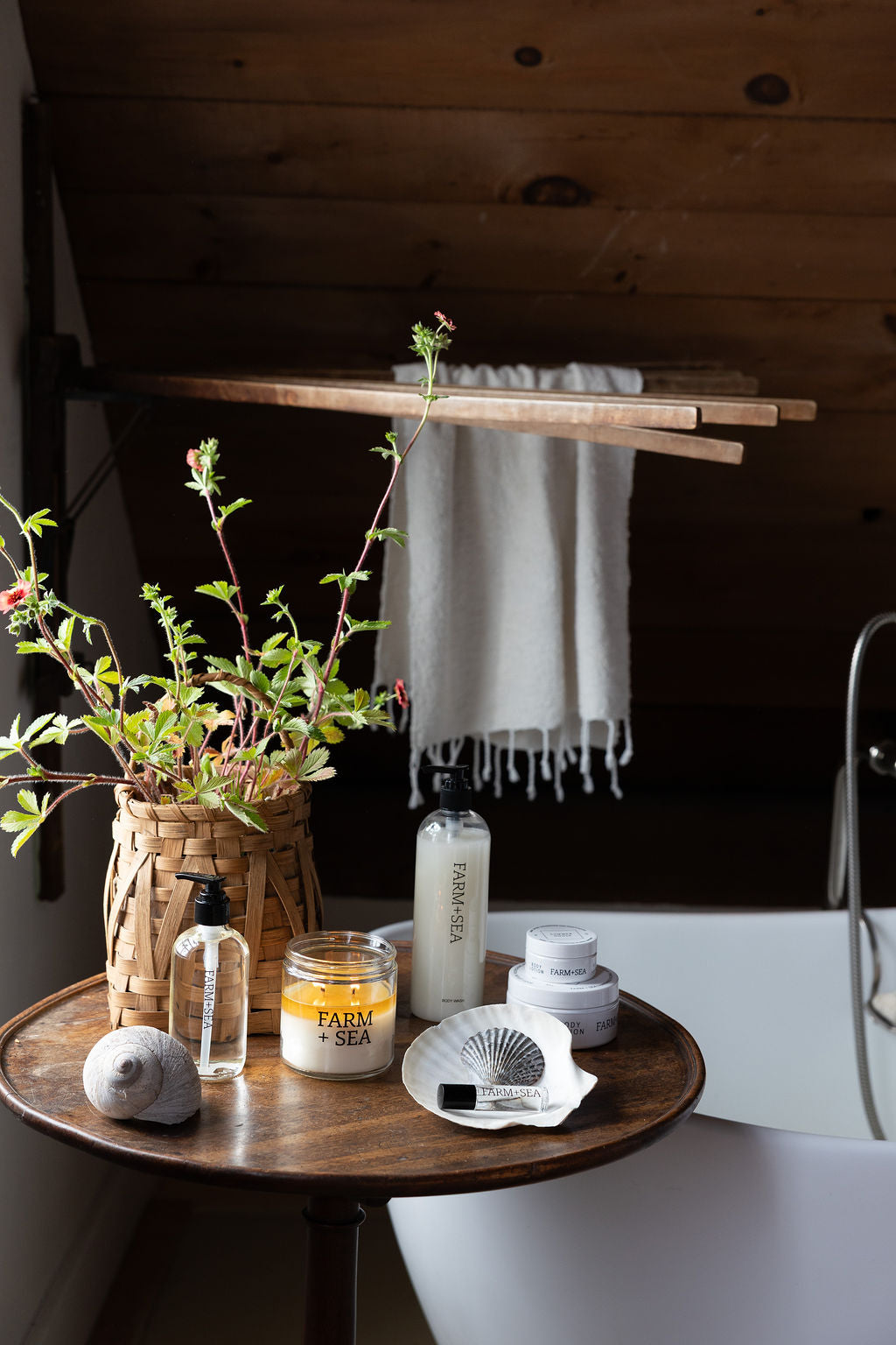 A bathroom scene with a round wooden side table holding a woven basket planter with green stems and Farm + Sea bath and body products: body oil, body lotion, body wash, and roll-on perfume. A white towel hangs from a wooden rack on the dark wood wall behind, and part of a white bathtub is visible.