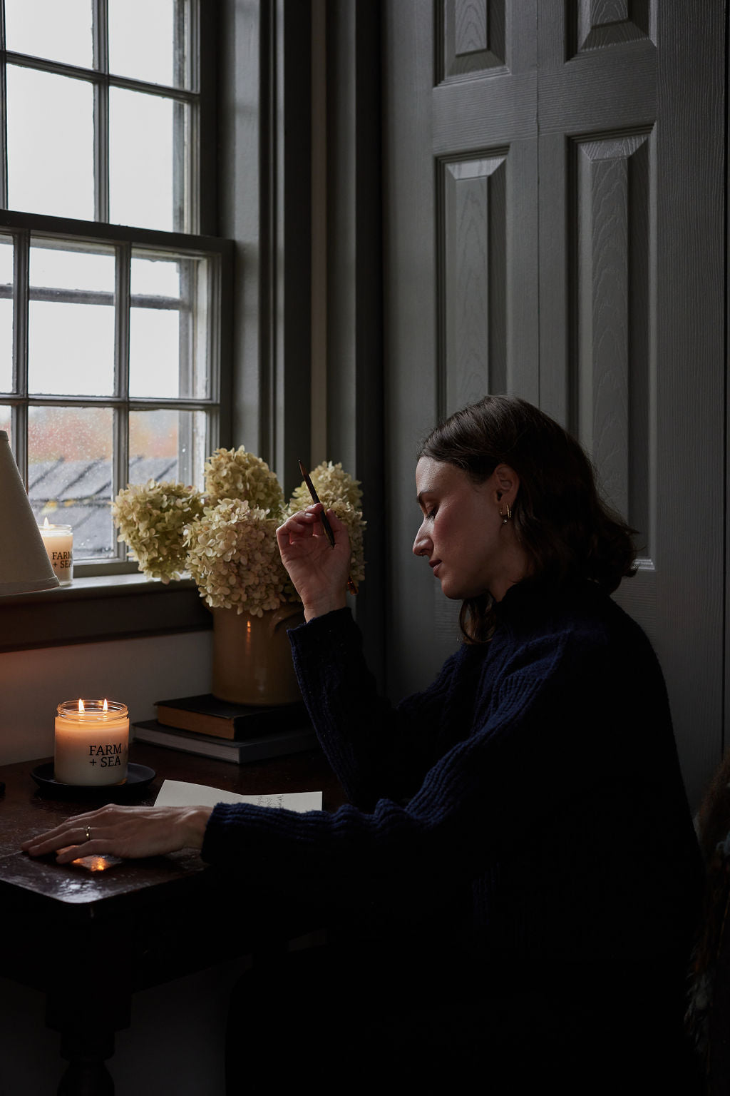 A woman in a dark navy knit sweater sits at a wooden desk by a window, holding a pen. A lit Farm + Sea candle is visible on the desk next to a vase of pale hydrangeas and a dark paneled door.
