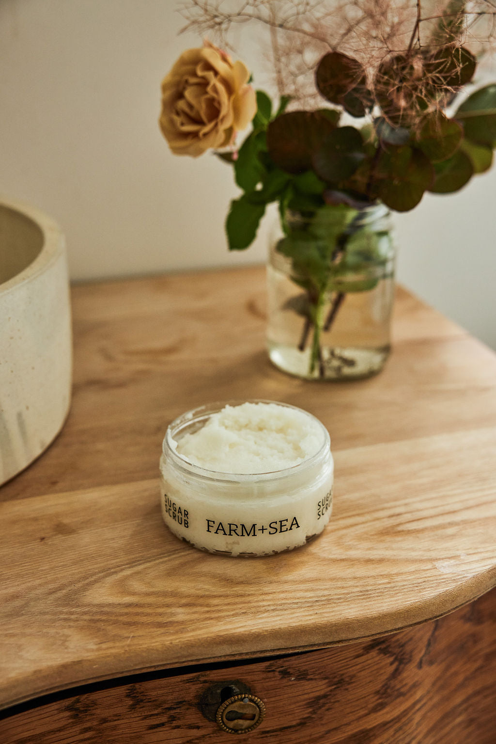 Jar of Farm + Sea sugar scrub on a wooden surface with flowers in the background
