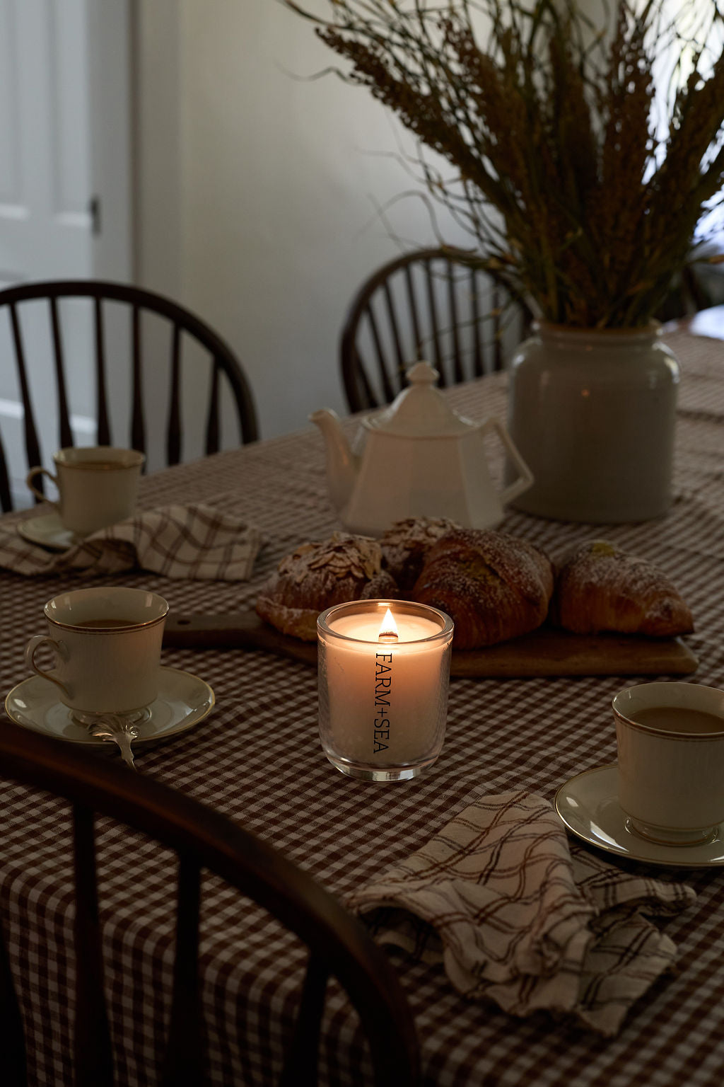 A lit glass jar candle with a white label reading 'FARM + SEA' sits on a brown-and-white checkered tablecloth, surrounded by teacups, bread, a white teapot, and a large vase of dried grasses in a dining room setting.