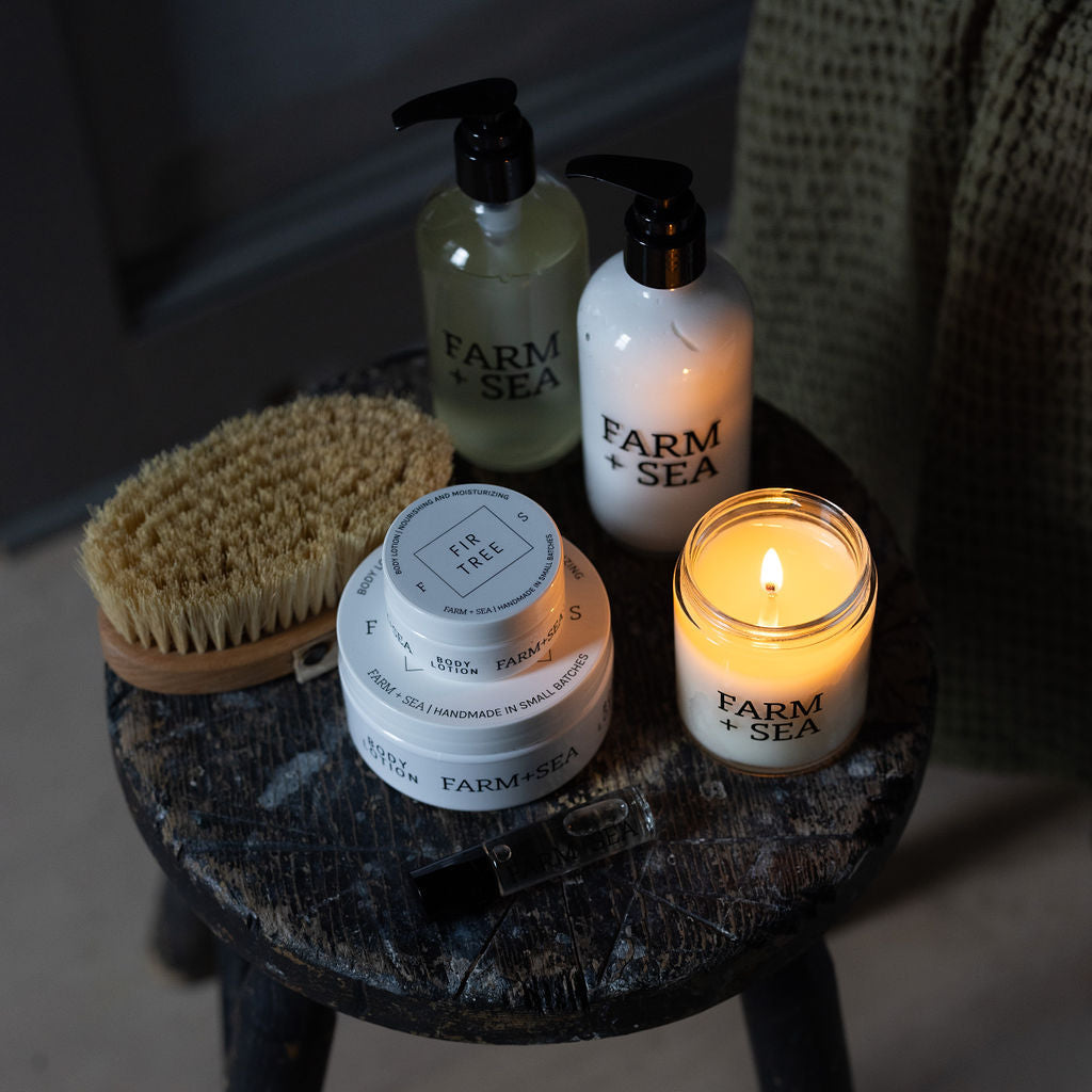 A Farm & Sea bath and body set arranged on a dark wooden stool, including body lotion, liquid hand soap, a classic candle, and a natural hair brush, photographed in a dimly lit indoor setting.