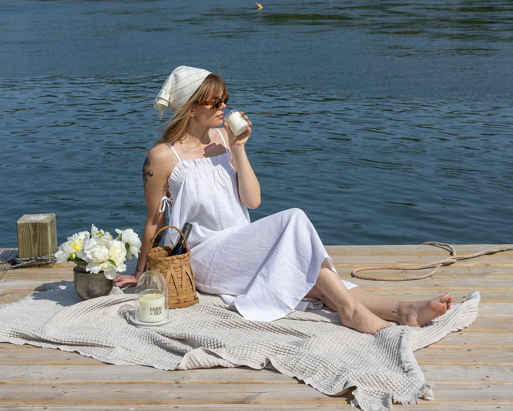 Woman in a white dress sitting on a dock by a lake, enjoying the smell of a Farm + Sea candle