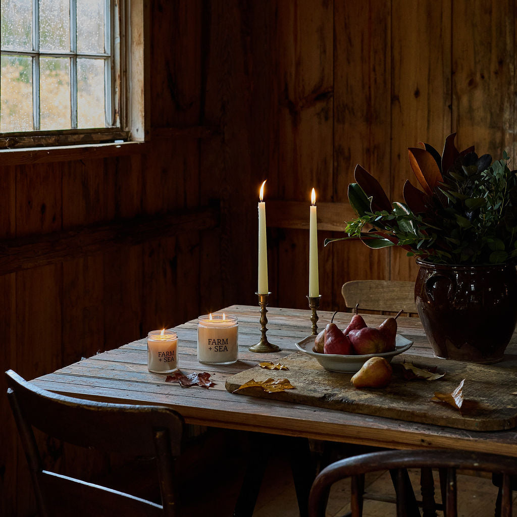 A rustic wooden dining table in a barn interior with two lit taper candles, the large and small Farm + Sea Classic Candles, a bowl of red pears, dried leaves, and a potted plant. 