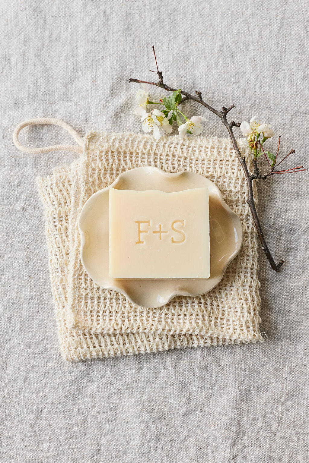 Bar of Farm + Sea soap on a textured surface with a mesh soap saver and branch with flowers.