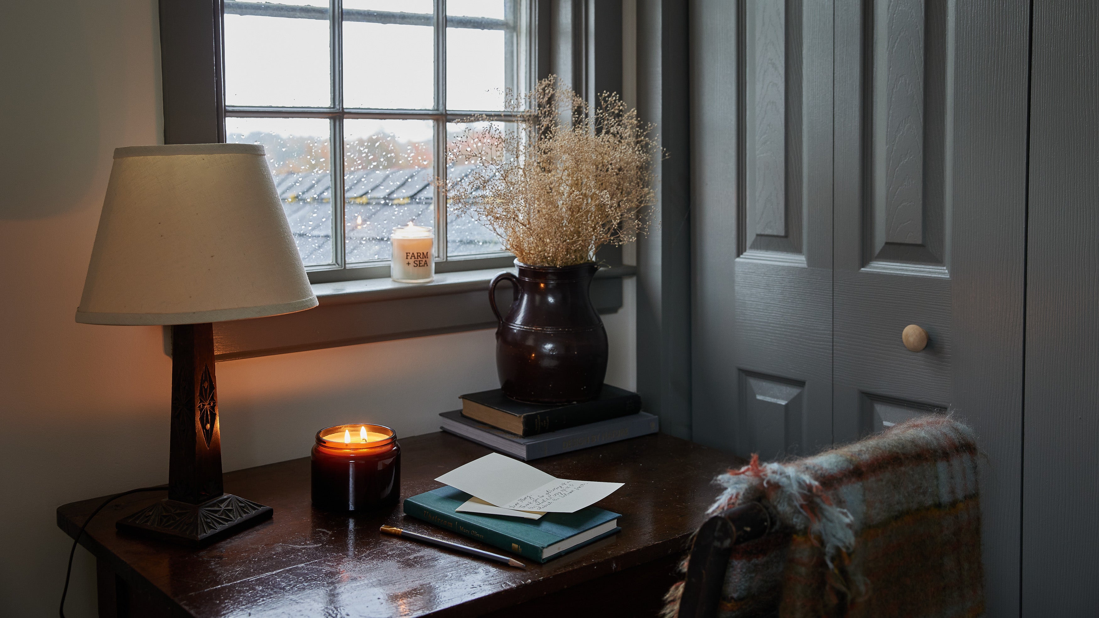 A cozy interior scene with a dark wooden desk by a window, lit by a table lamp and Farm + Sea candles. A ceramic vase with dried flowers sits on stacked books, and papers rest on the desk.