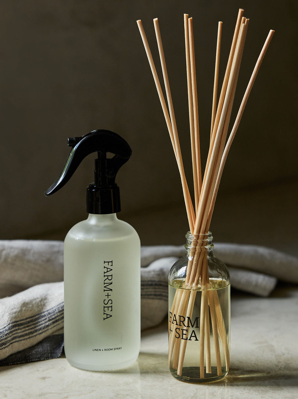Farm + Sea Room + Linen Spray and a reed diffuser on a marble countertop with a towel in the backgroud