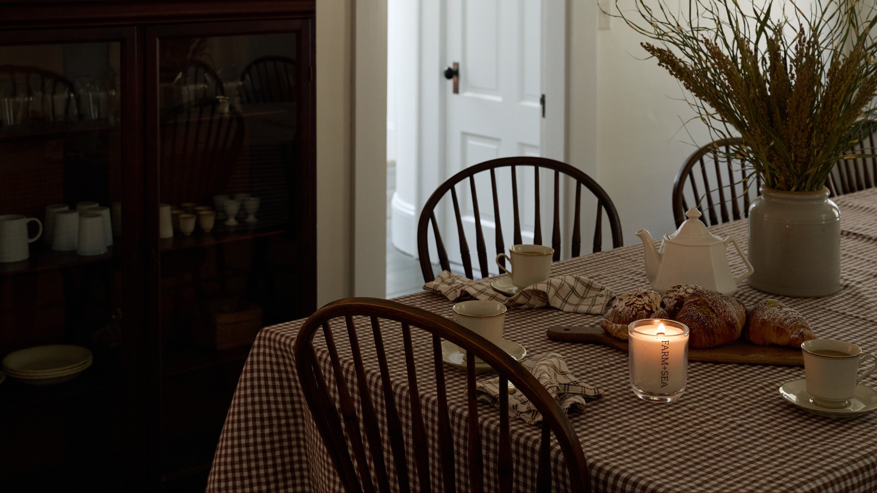 A cozy dining room scene with a checkered tablecloth, wooden spindle-back chairs, a lit Farm + Sea a Tumbler Candle, bread, cups, and a large vase of dried branches. A dark wood hutch stands on the left, and a white door is in the background.
