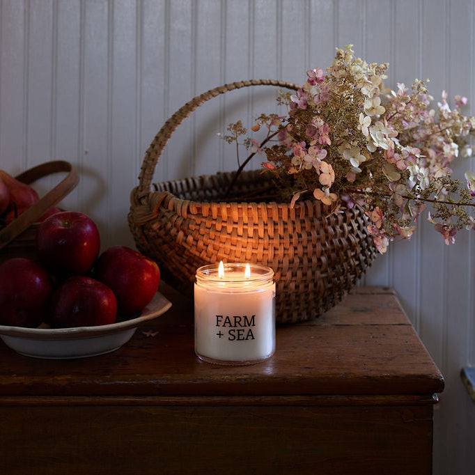 A lit white candle labeled 'FARM + SEA' sits on a wooden table beside a woven basket with dried flowers, a bowl of red apples, and another basket of pears against a paneled wall.