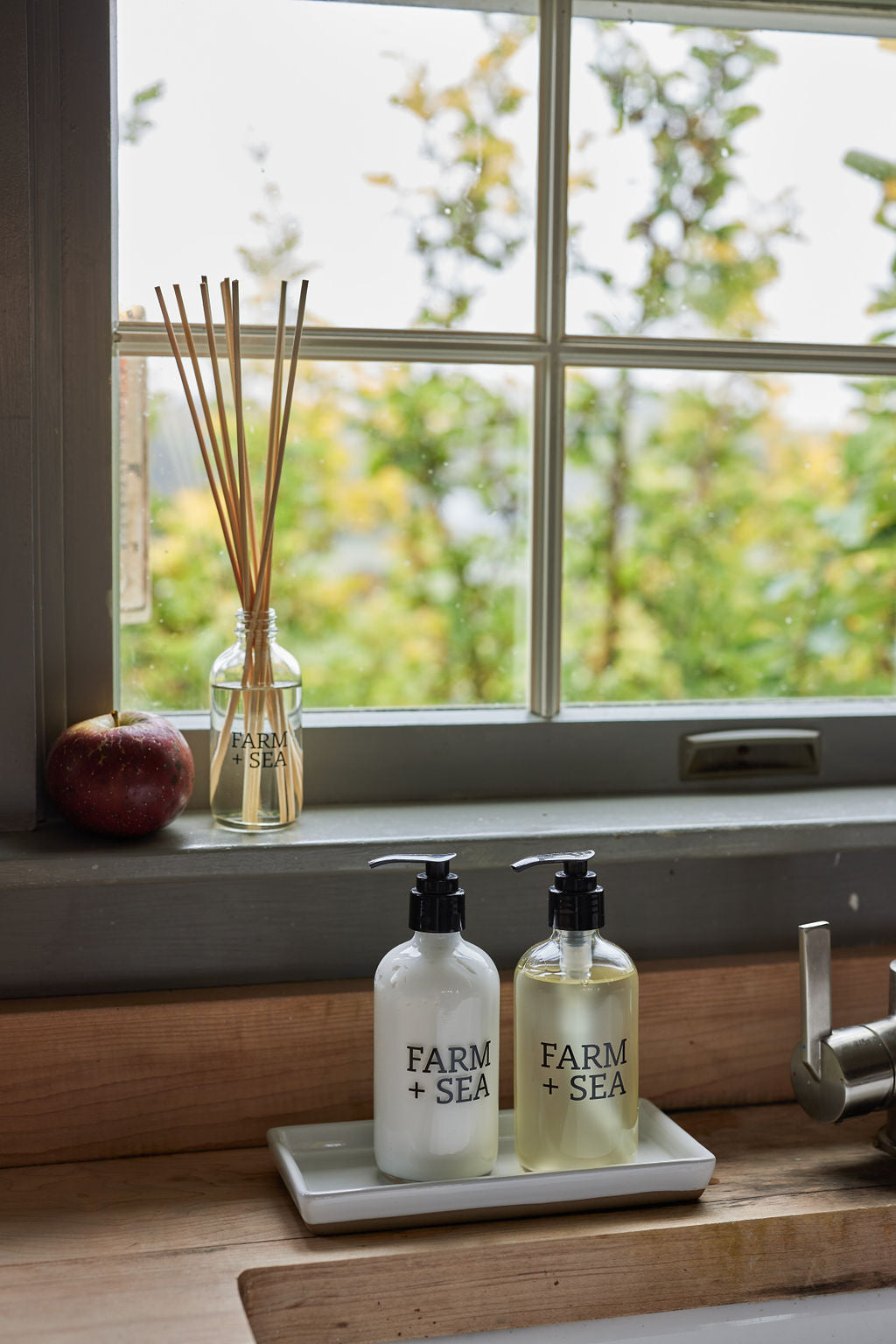 Two Farm + Sea pump bottles on a small tray in a sink area, with a window behind them showing greenery outside. A Farm + Sea reed diffuser and a red apple sit on the windowsill.