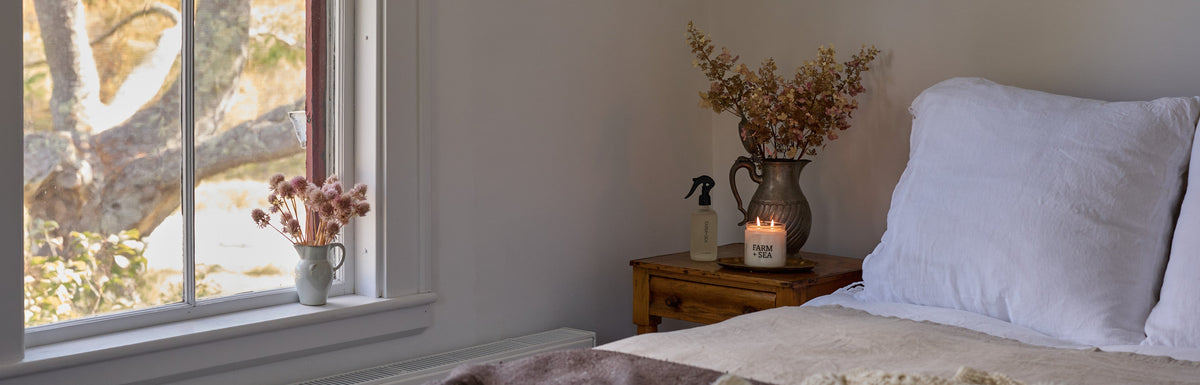 A cozy fall bedroom scene with a neatly made bed featuring white pillows, a cream knit throw blanket, and a dark brown textured sheet. A wooden nightstand with a vase of dried flowers and a lit Farm + Sea classic candle and Room + Linen spray sit beside a window with a view of trees outside.