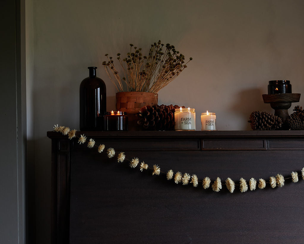 A dark vintage upright piano in a dimly lit room, decorated with a beaded garland, Farm + Sea Classic and Sugar Shack Candles, pinecones, and dried flowers on top, with a rustic wooden bench in front.