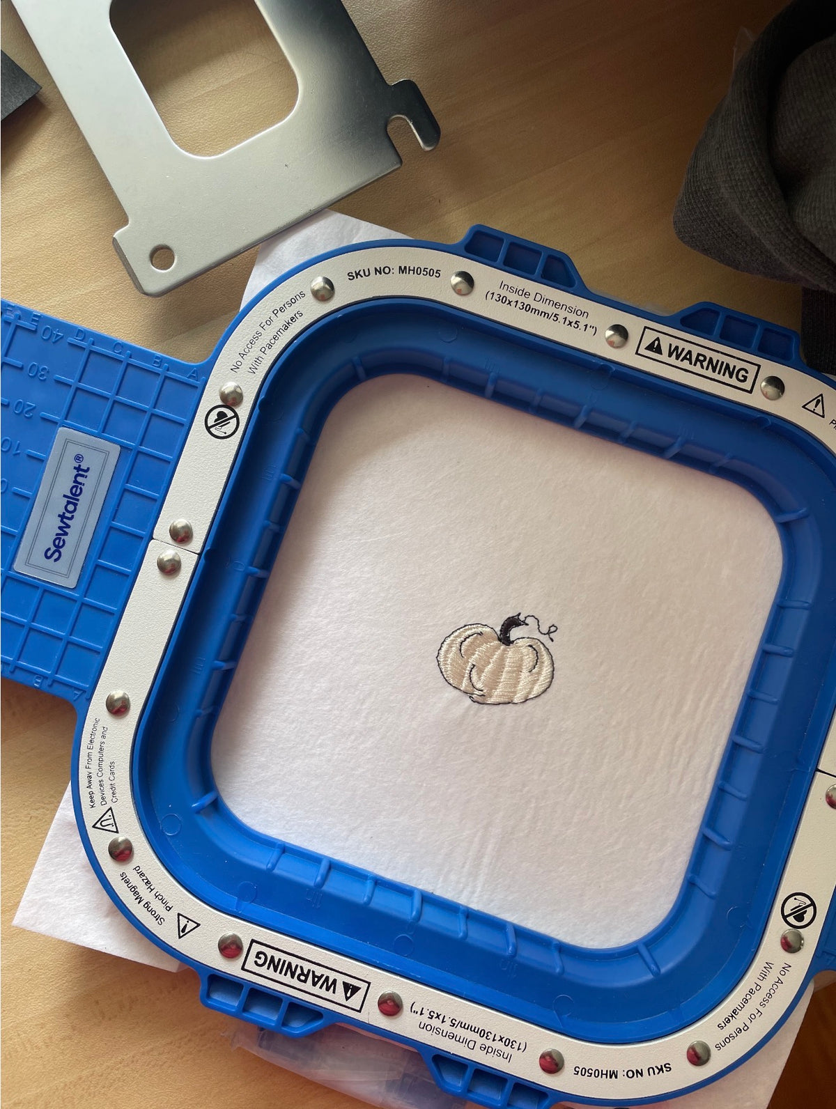 A close-up of a blue and white embroidery machine frame with a white fabric patch containing a small embroidered white pumpkin design. A blue ServaVolt control pad is visible on the left, and a metal mounting bracket is partially visible at the top on a wooden surface.