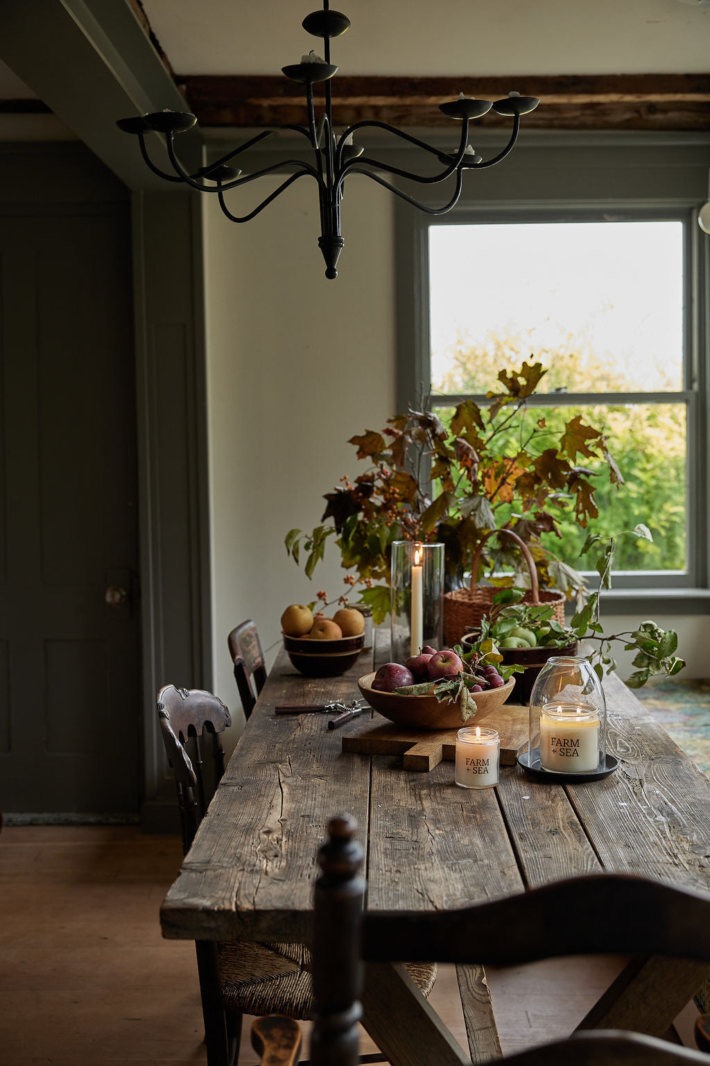 A rustic wooden dining table set with Farm + Sea candles, bowls of fruit, and potted plants in a cozy room with a window, chandelier, and dark wooden chairs.
