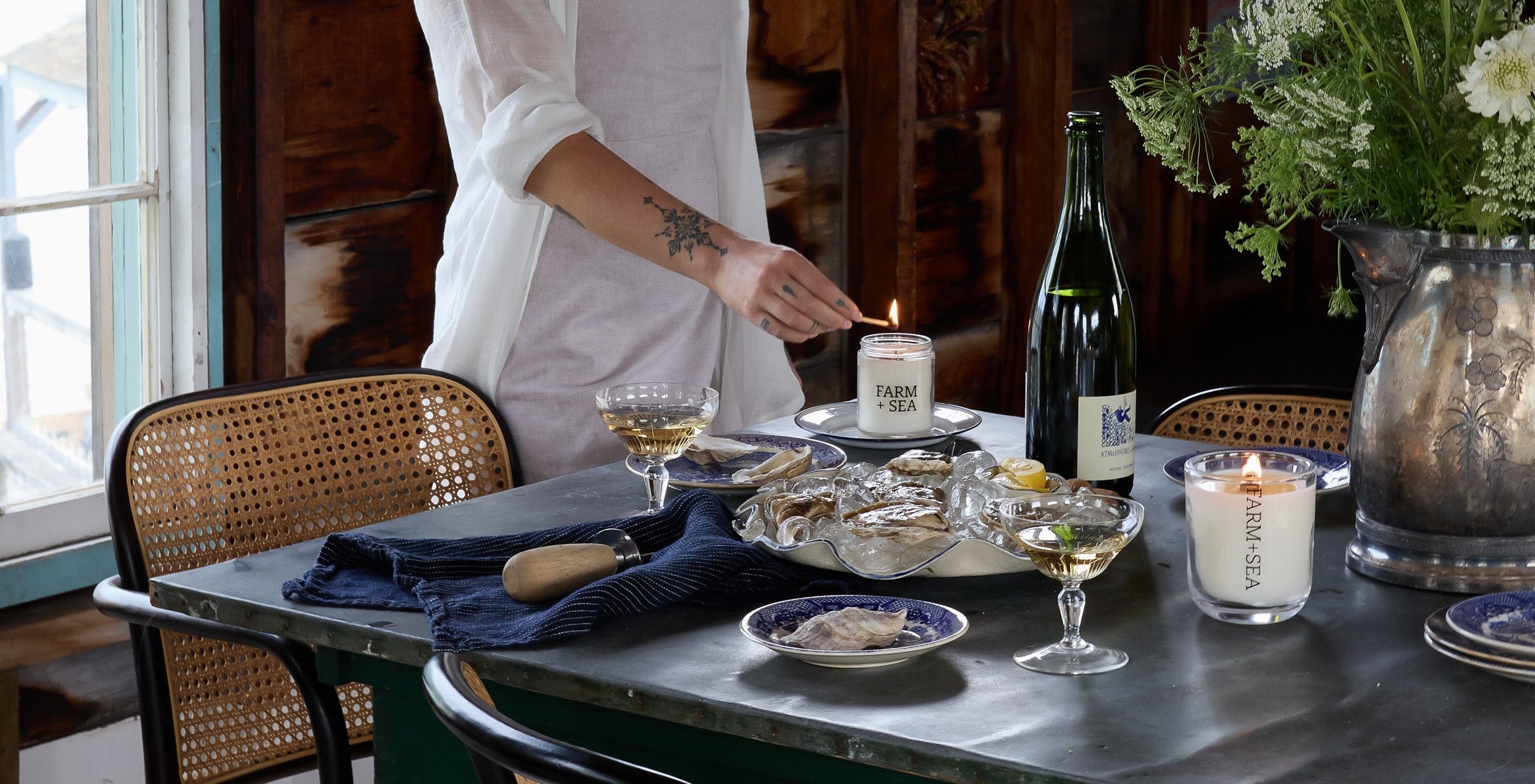 A woman in a white outfit lighting a Farm + Sea classic candle at a table set for a meal in Maine with oysters and wine.