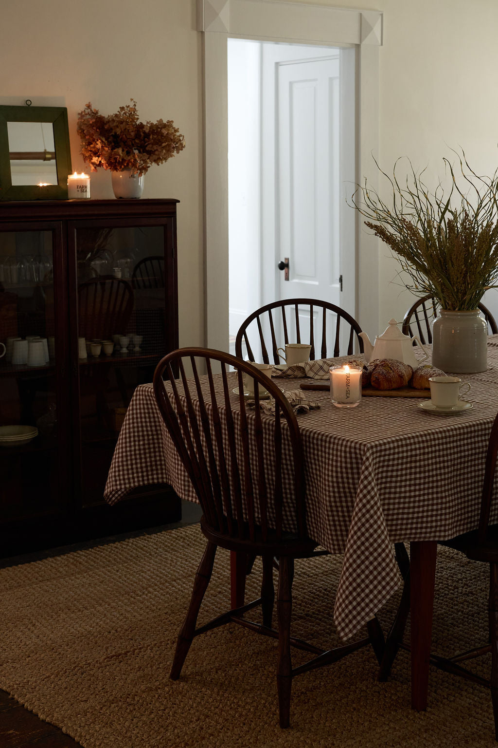 A dining room scene with a two Farm + Sea candles, checkered tablecloth on a round table, dark wooden chairs, candles, and dried floral arrangements in a vase, set in a home interior with a cabinet and open doorway in the background.